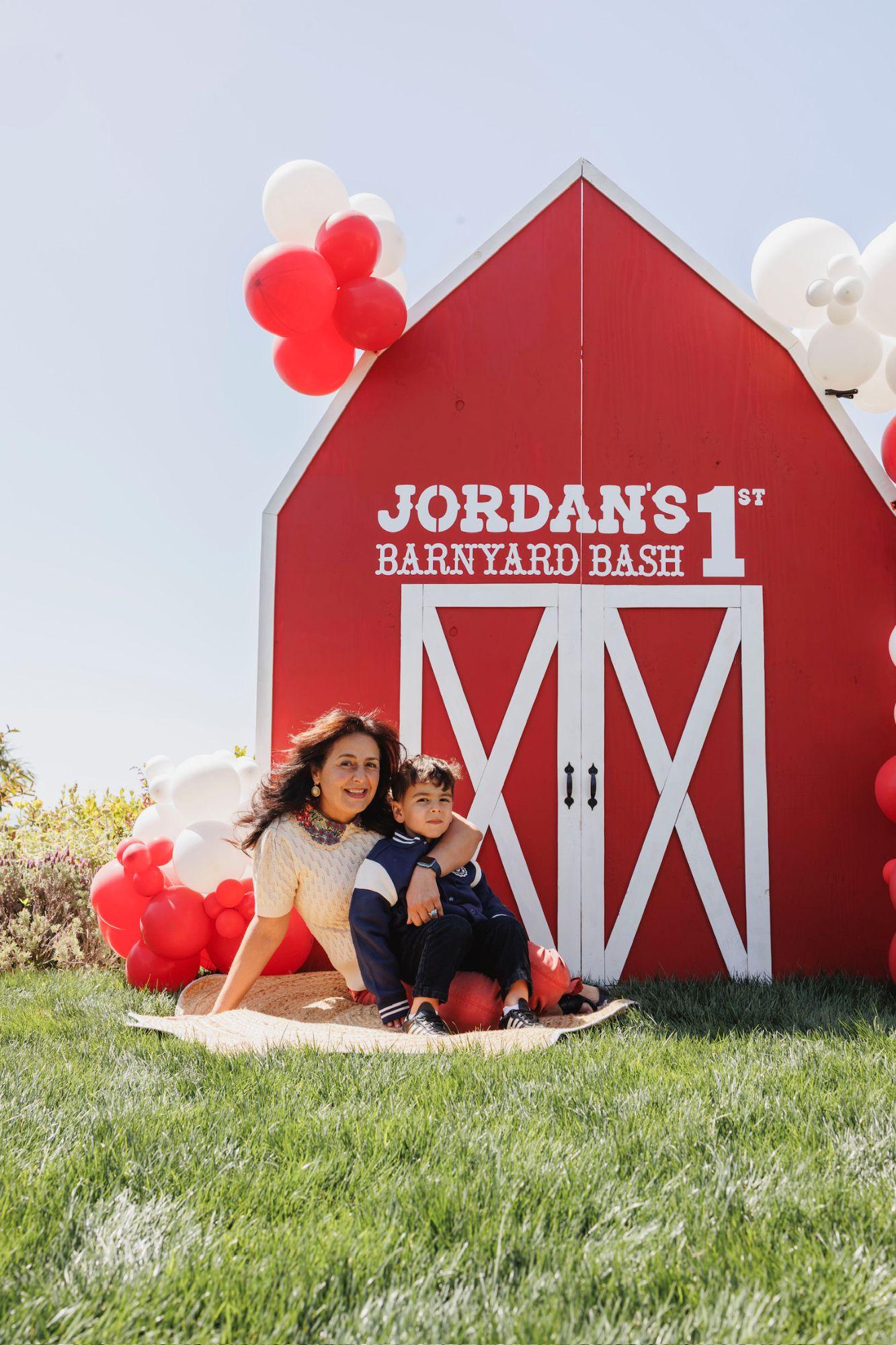 A red barn backdrop with a first birthday sign and red and white balloons, with a woman and a small boy sitting on a rug in front of it