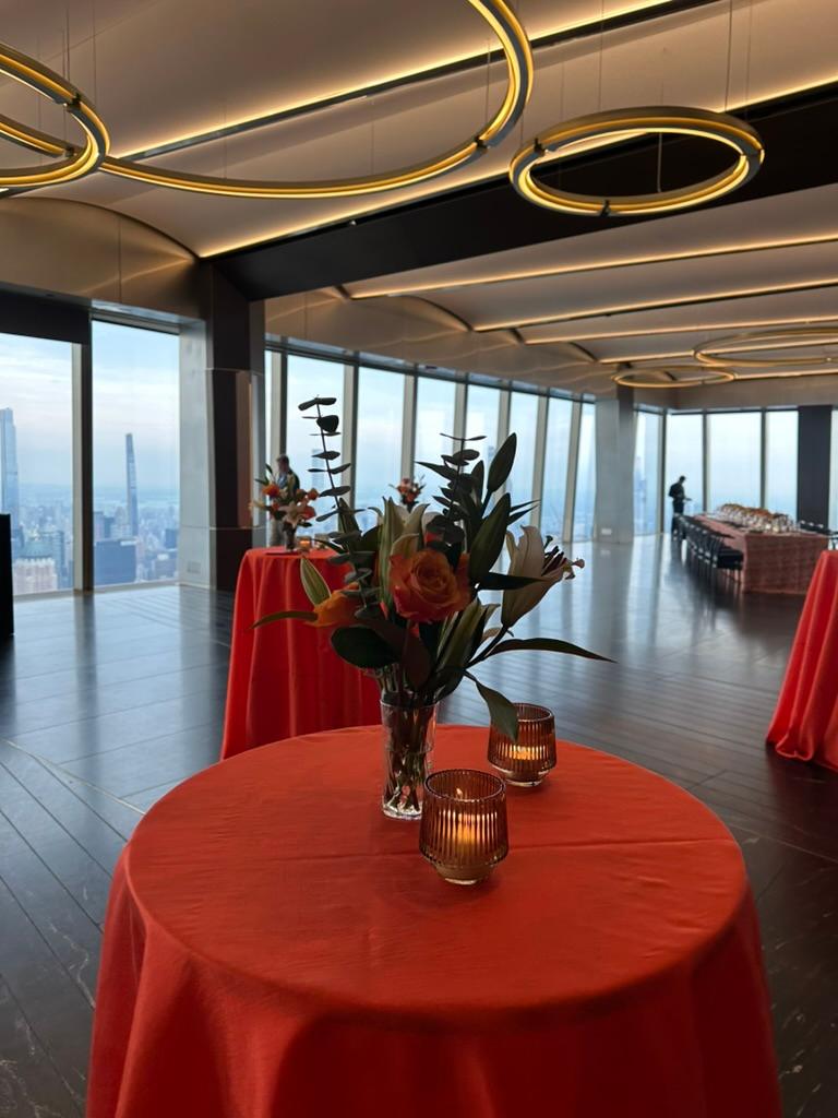 Cocktail tables in orange linen with candles and a small arrangement of roses and lilies, in a high-floor event space with floor-to-ceiling windows over the Manhattan skyline