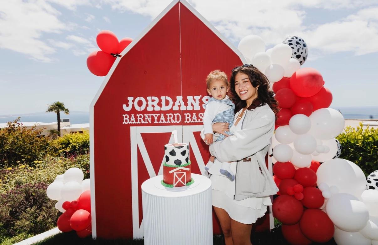 Debby holding the birthday boy in front of the red barn backdrop and the first birthday cake, framed by red, white and cow-print balloons