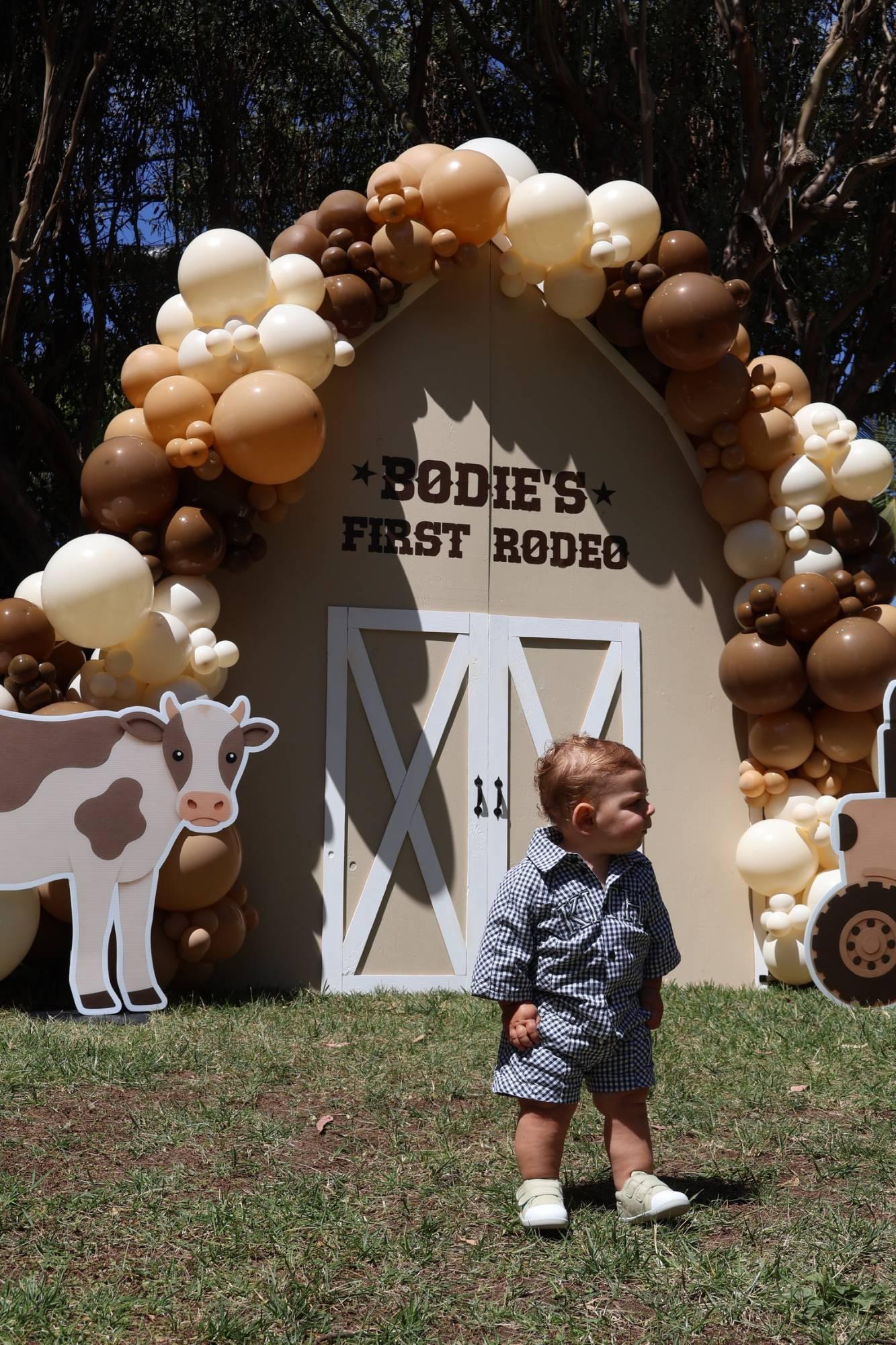 A small boy in a gingham outfit in front of a cream barn backdrop framed by brown and cream balloons, beside a cow cutout