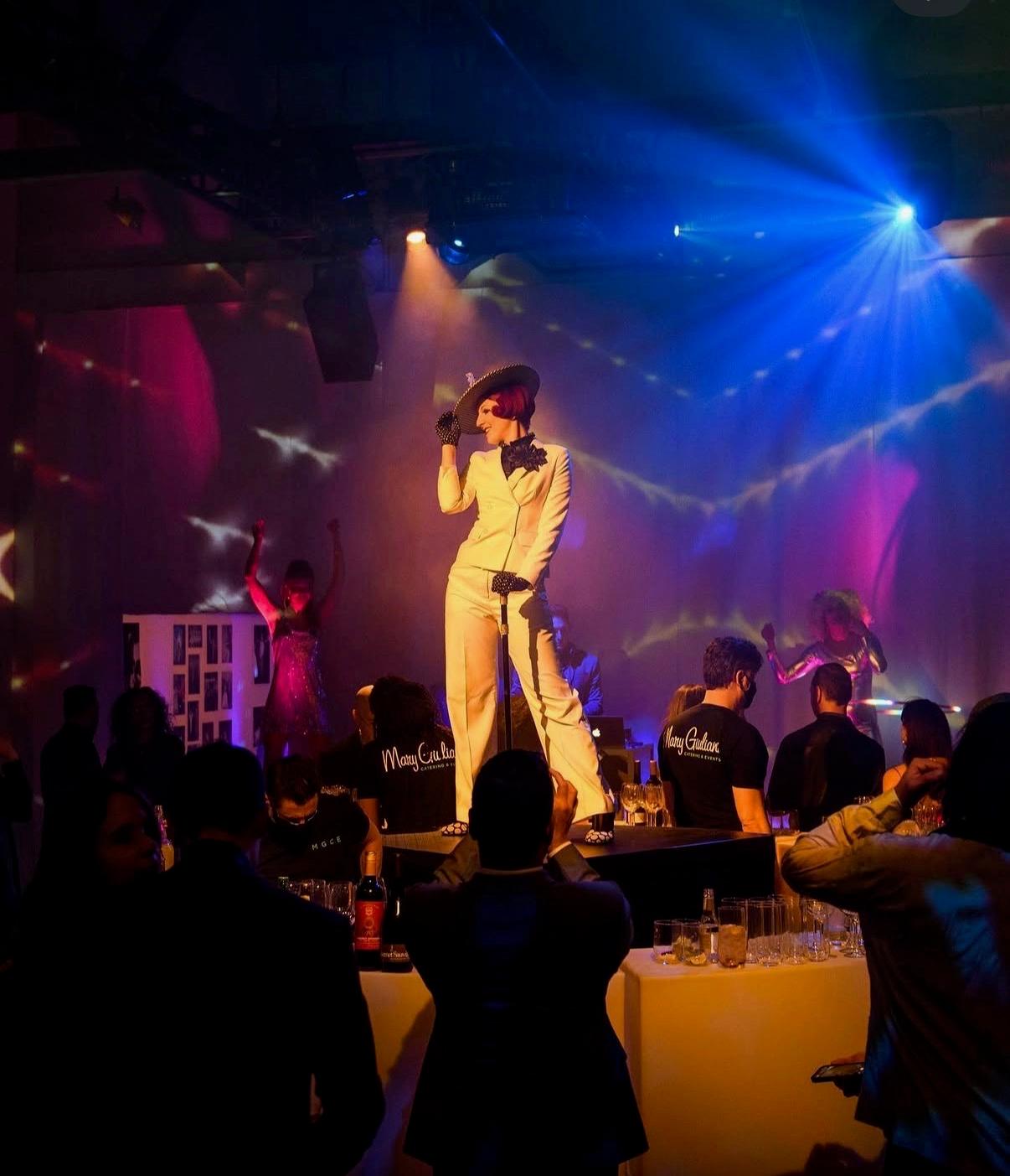 A performer in a white suit and wide hat on a small stage under blue and pink lights, with dancers behind her and guests watching from the floor