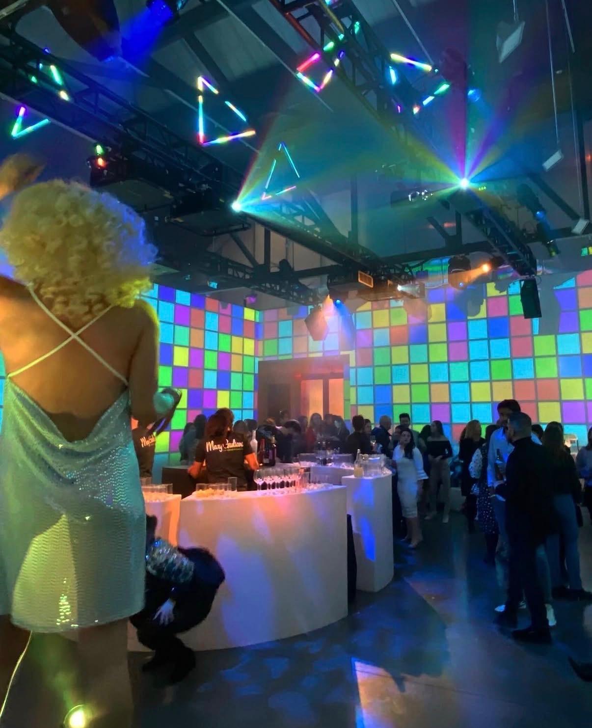 A performer in a sequined slip dress and a blonde curly wig beside a glowing white bar, with walls of projected colored squares and neon triangles overhead