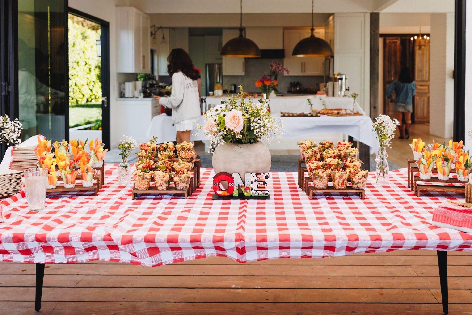 A buffet table under a red gingham cloth, with cups of pasta salad and vegetable crudités on wooden risers around a wildflower centerpiece
