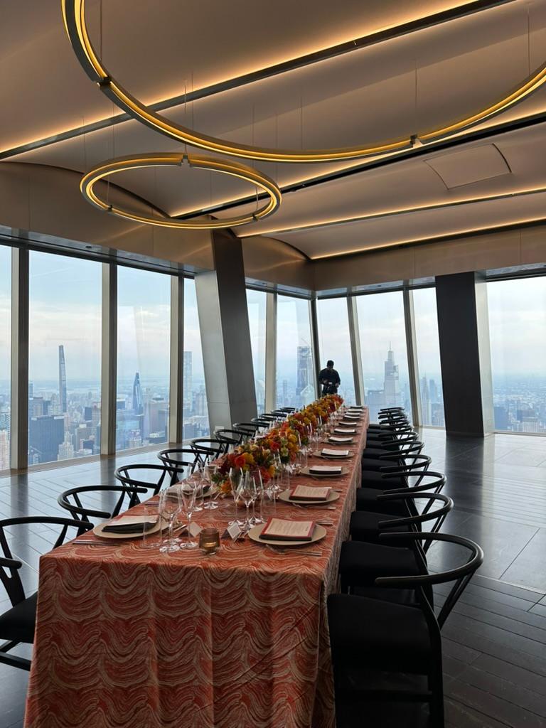 A long dinner table set for a seated dinner, with patterned orange linen, a low run of orange and yellow flowers, menus at each place and black wishbone chairs, beside windows over the Manhattan skyline