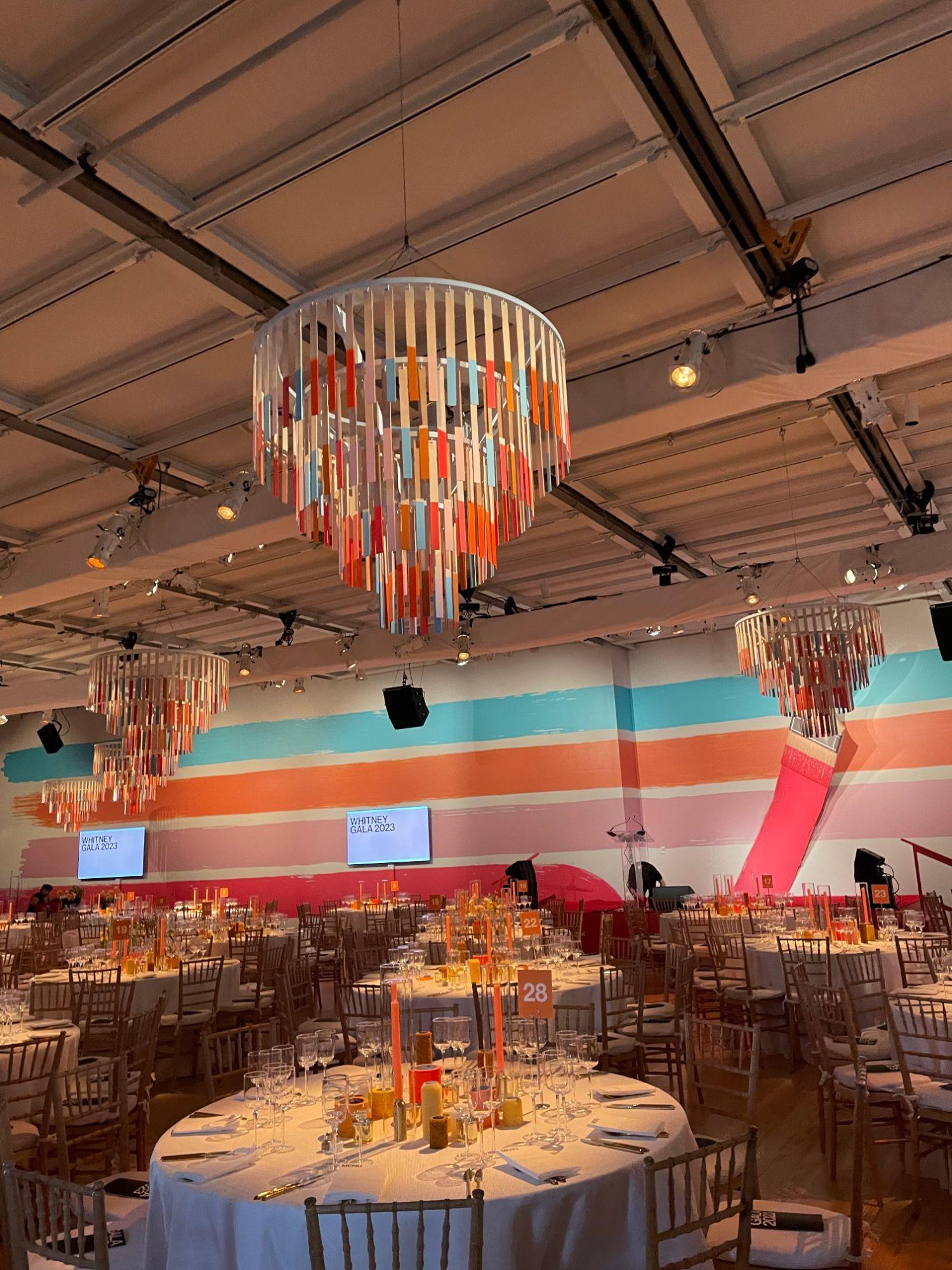 A gala dinner room set with round tables, tall orange candles and numbered table cards, under three tiered chandeliers of colorful painted strips, with broad painted stripes of blue, orange and pink across the walls