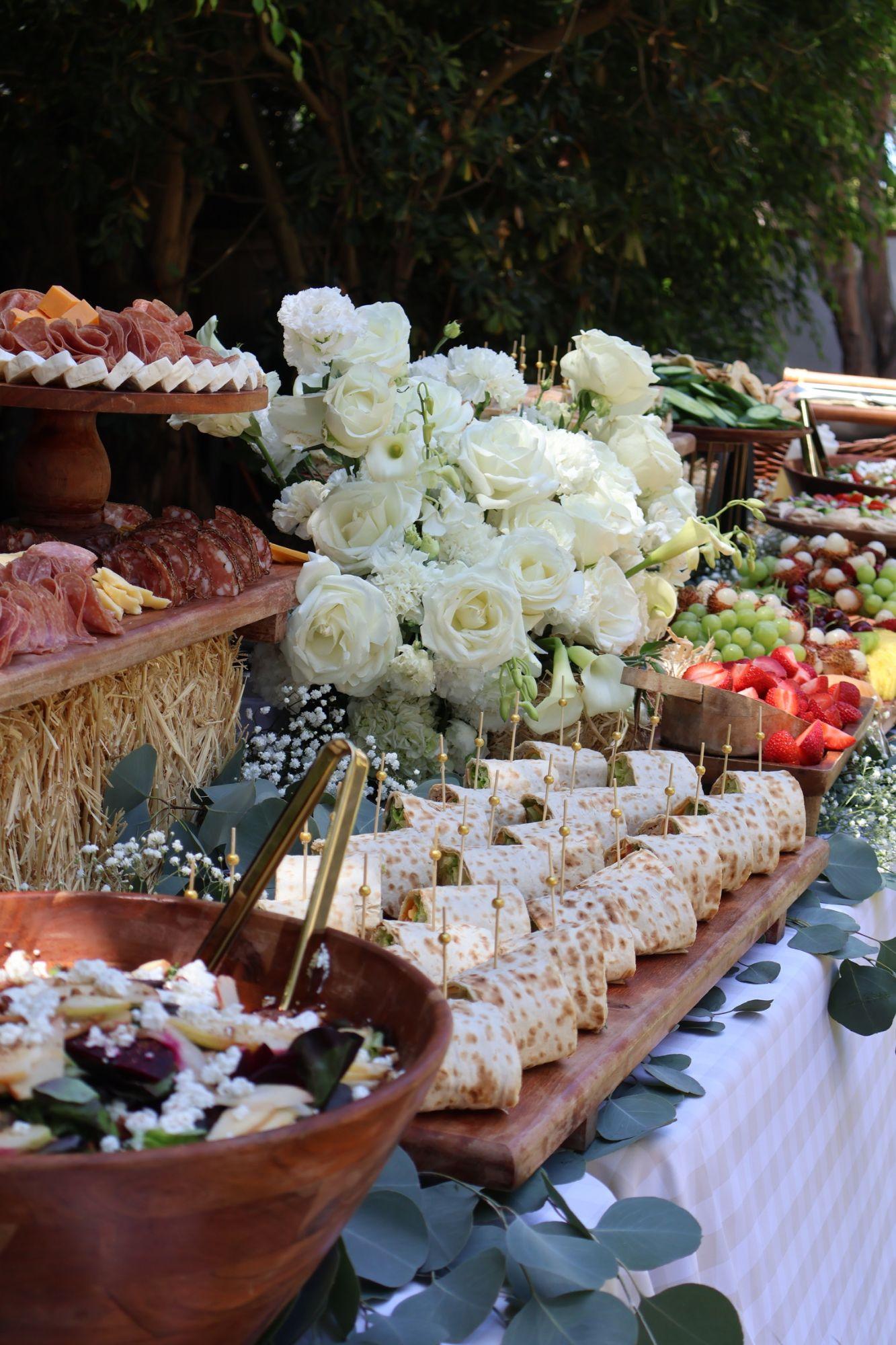 A grazing table of charcuterie, wraps, fruit and salad around an arrangement of white roses