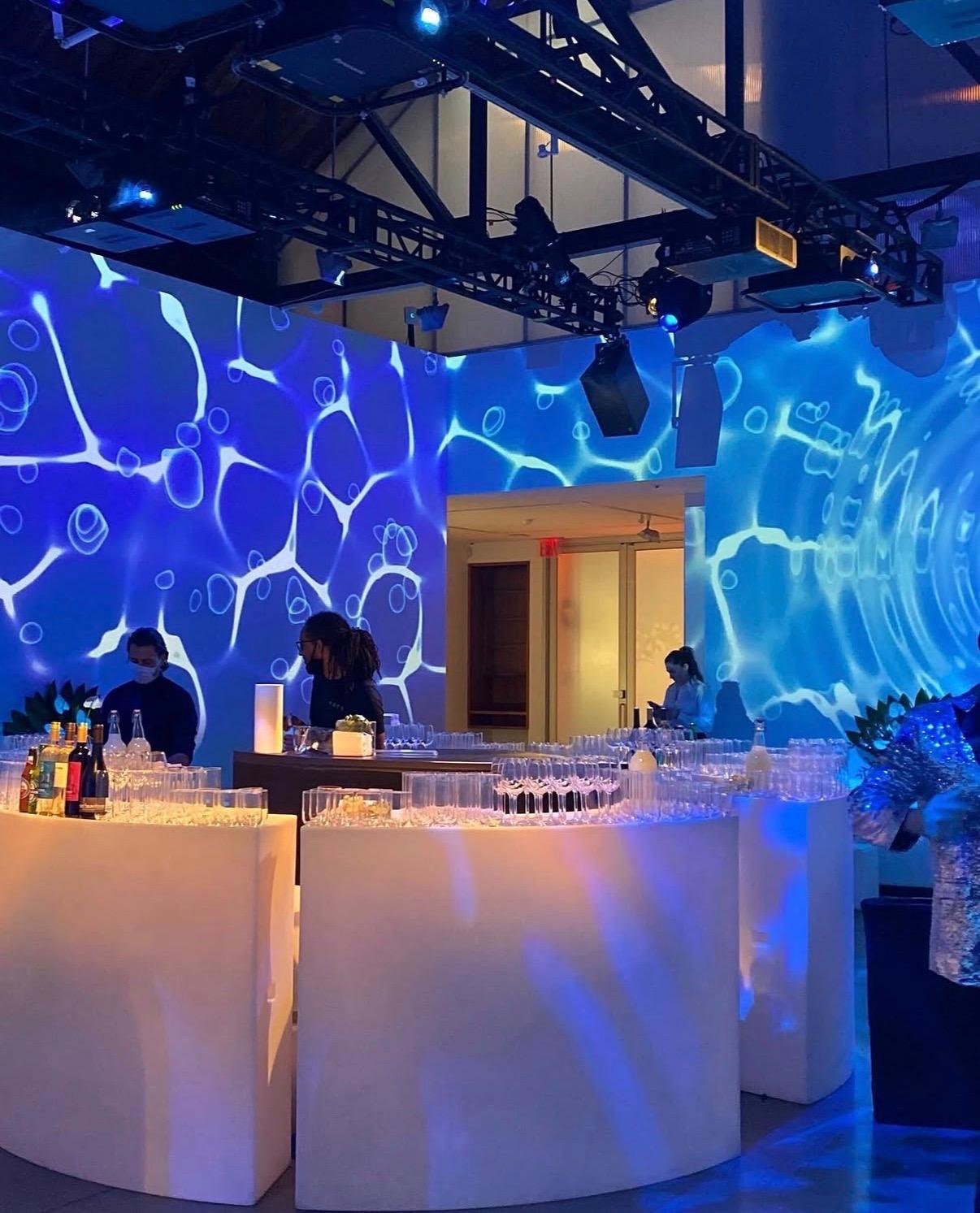 Curved white bars lined with glassware under walls projected with rippling blue water light