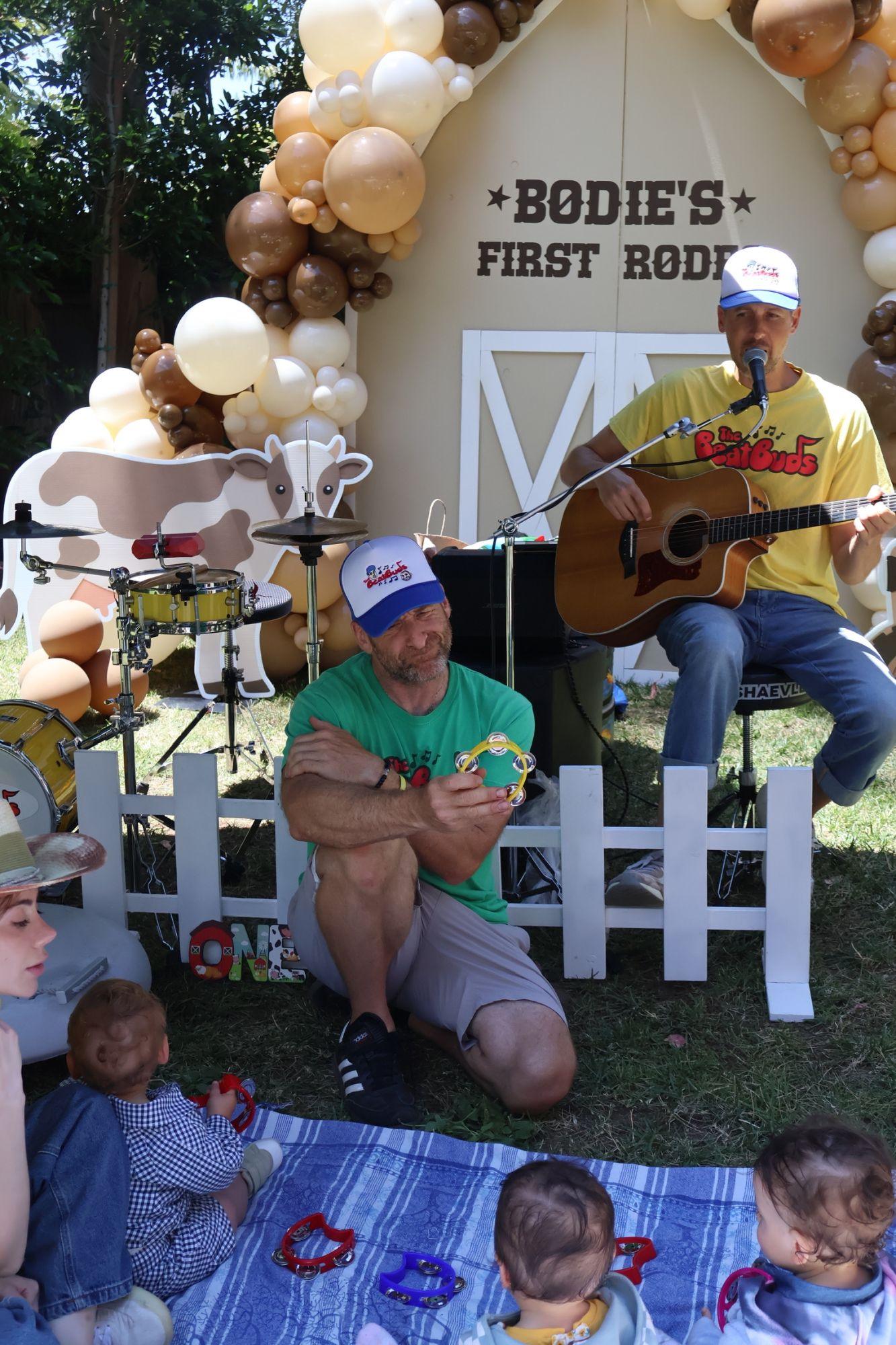 Two musicians with a guitar and a tambourine playing for toddlers on a picnic blanket in front of the barn backdrop