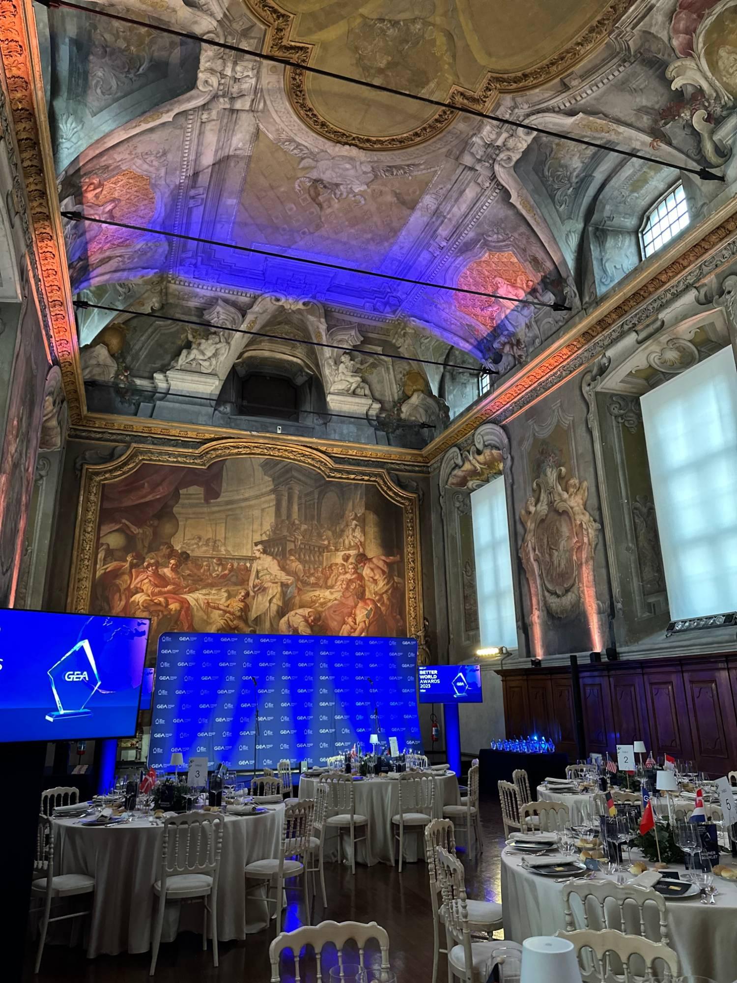 An awards dinner in a frescoed hall with a painted ceiling, round tables with white chairs and national flags, and a stage lit in blue