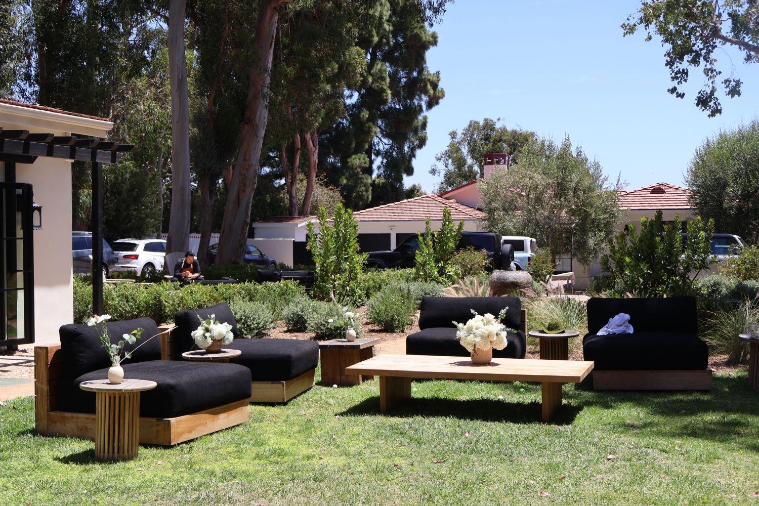 An outdoor lounge of black cushioned seating, teak tables and small white flower arrangements on a lawn
