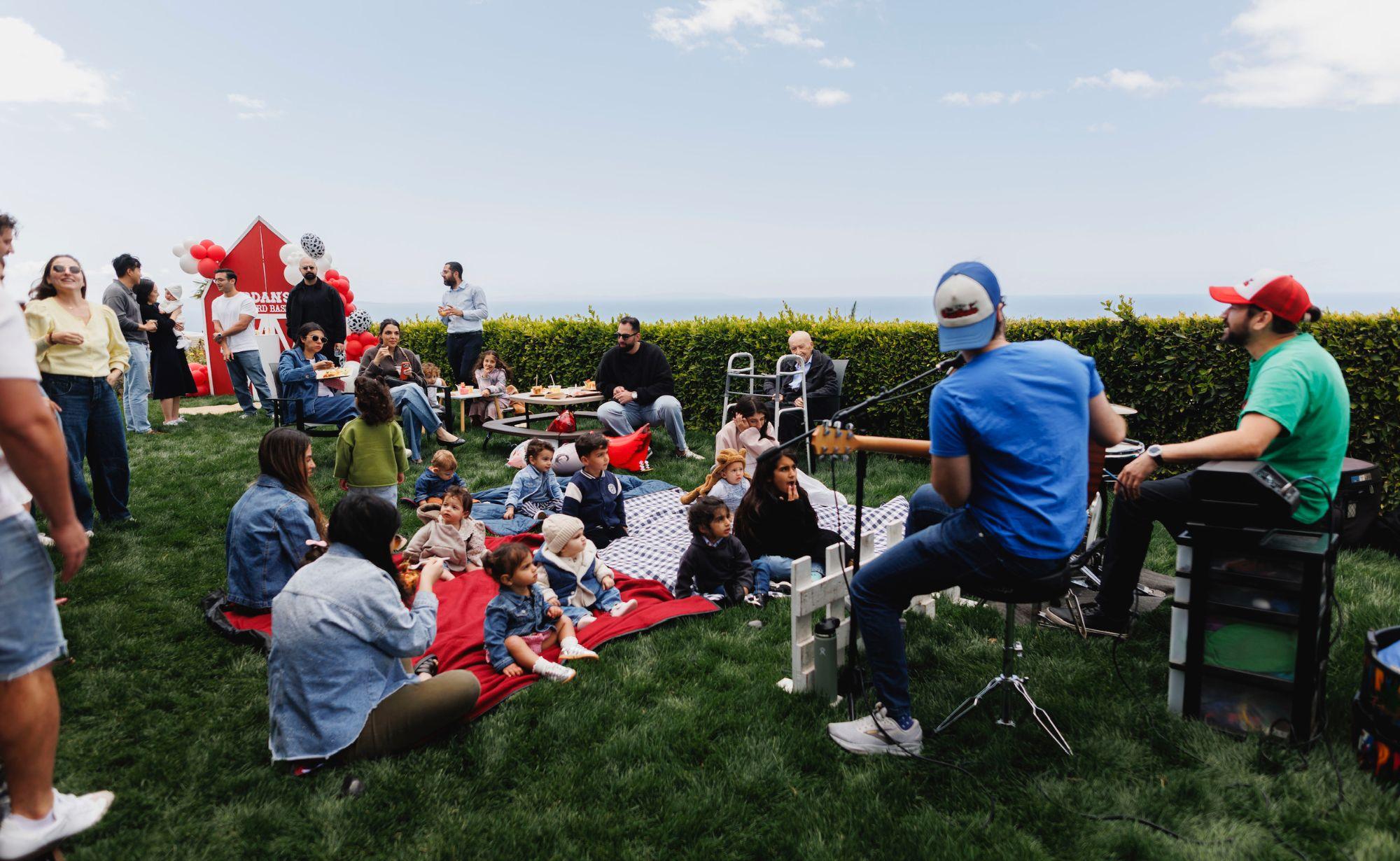 Families on picnic blankets on a lawn listening to two musicians, with the red barn and the ocean behind them